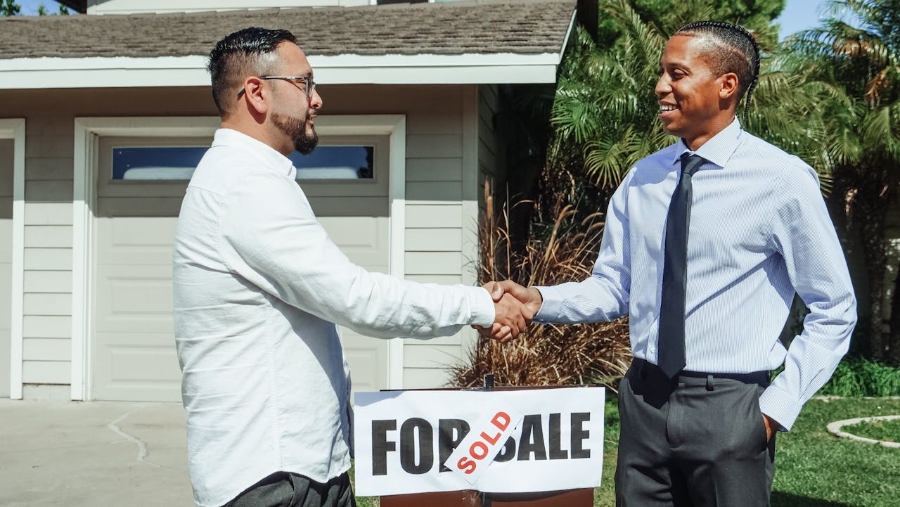 services-03 Two men shaking hands in front of house sold sign, sealing real estate deal outdoors.
